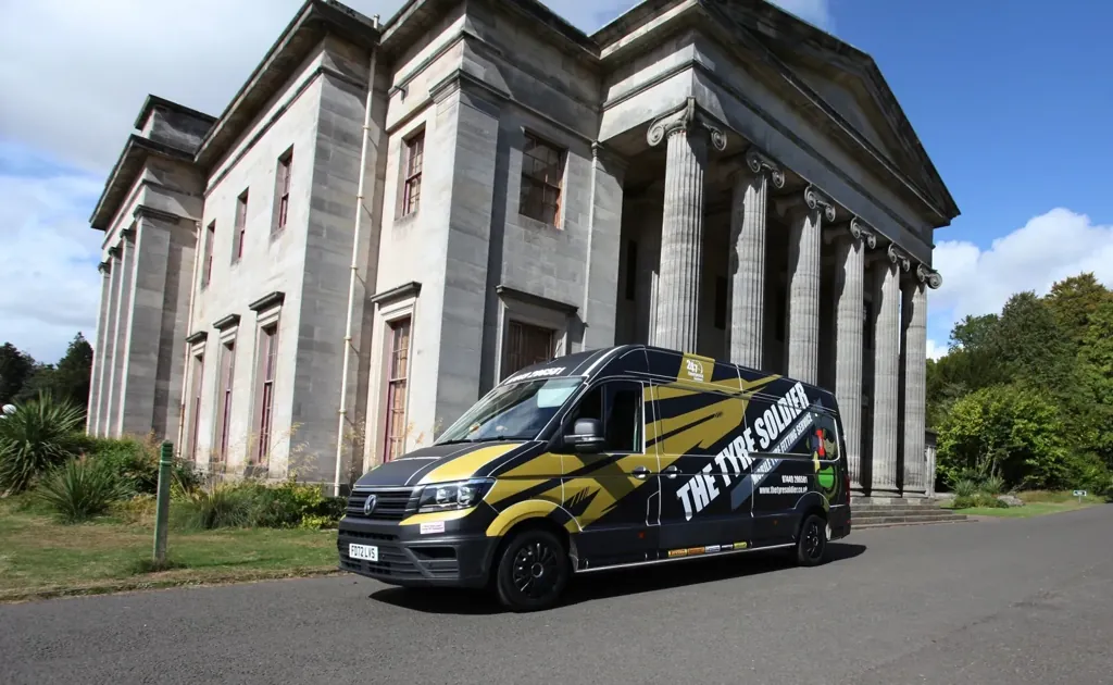 The Tyre Soldier black VW Crafter van — gold tribal-stripe livery, parked in front of a grand columned Georgian estate building at Camperdown, Dundee.